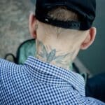 A man in a checkered shirt and hat showing a neck tattoo captured outdoors.