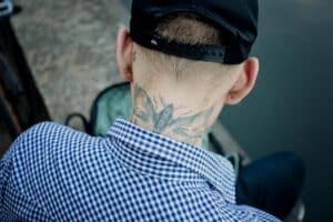 A man in a checkered shirt and hat showing a neck tattoo captured outdoors.