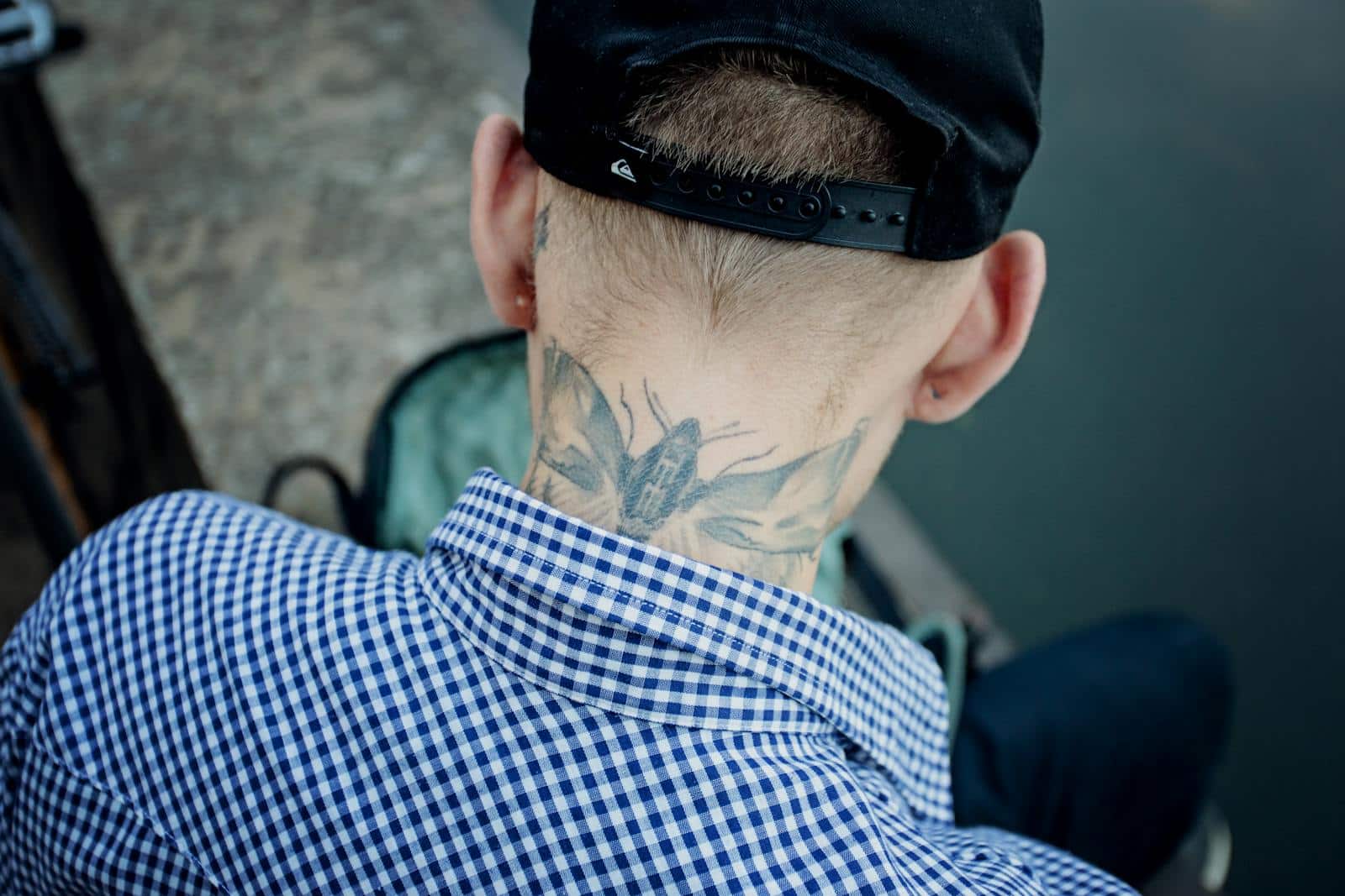 A man in a checkered shirt and hat showing a neck tattoo captured outdoors.