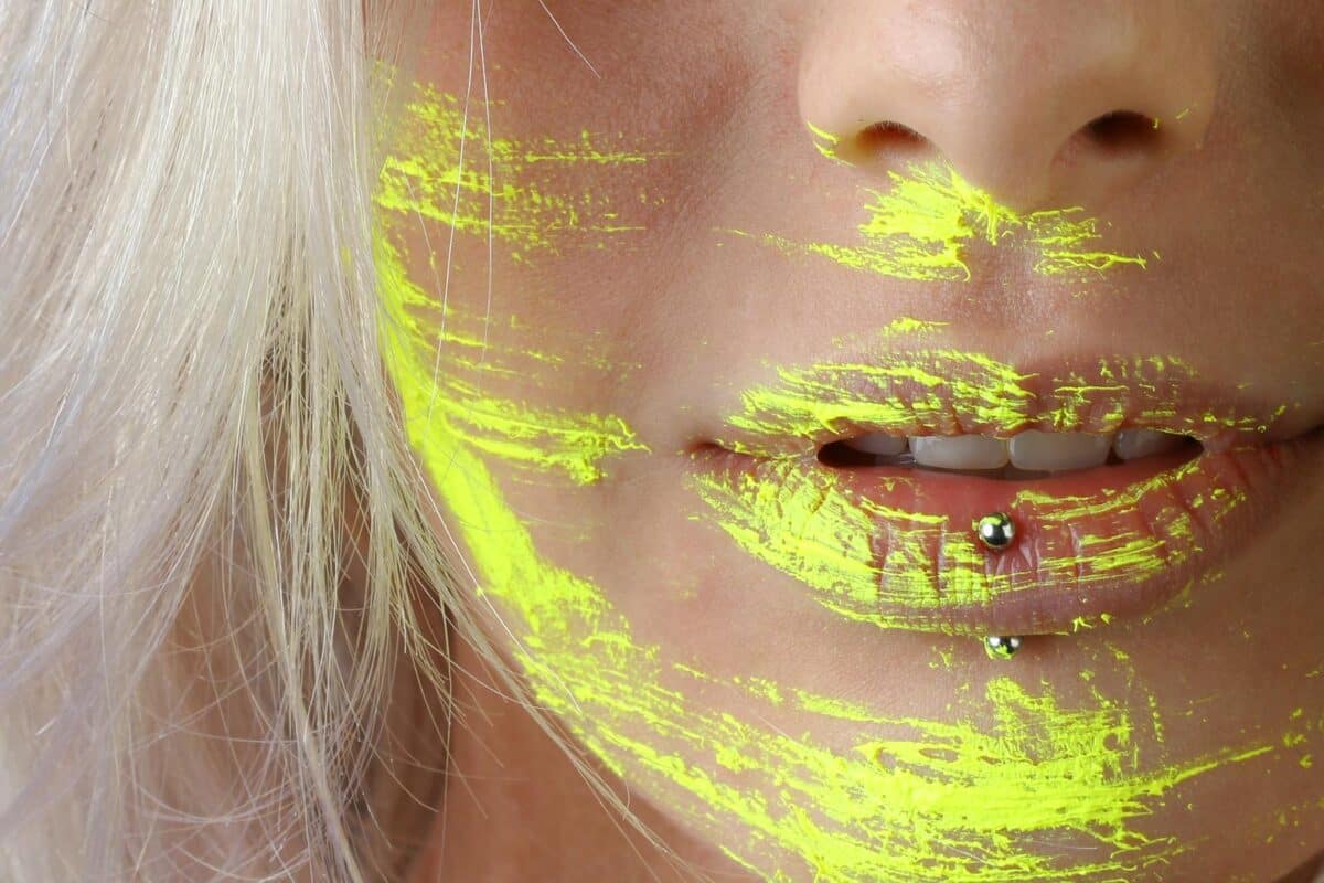 Artistic close-up photo of woman's lips with neon yellow paint and piercing.