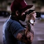 A tattooed photographer in a cap capturing a street scene at night.