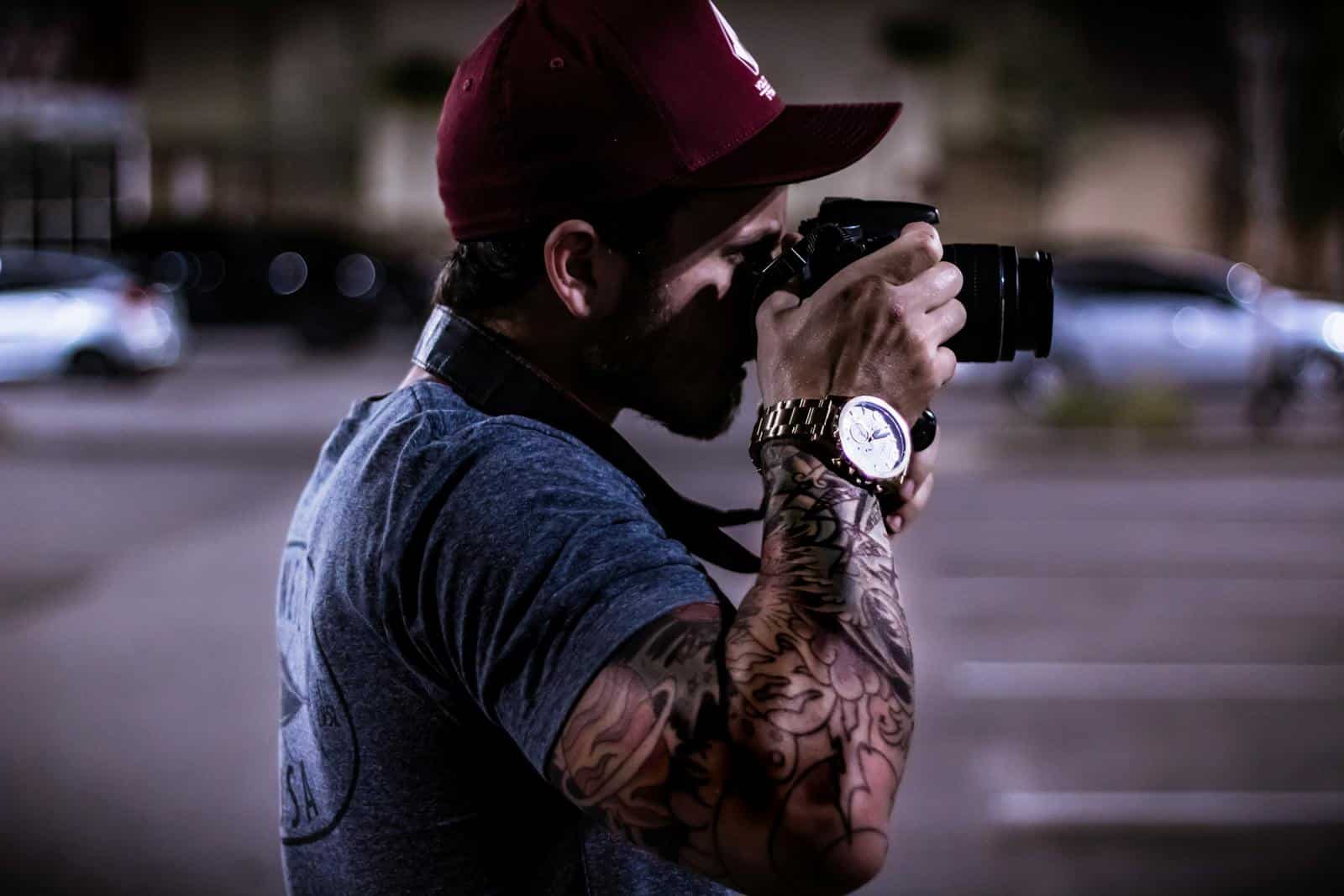 A tattooed photographer in a cap capturing a street scene at night.