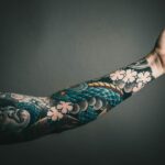 A close-up of a tattooed arm with a colorful sleeve design, featuring floral and artistic elements.