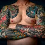 Close-up of a shirtless man with colorful sleeve tattoos and chest art.
