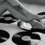 Black and white photo featuring tattooed legs surrounded by vinyl records on a bed.