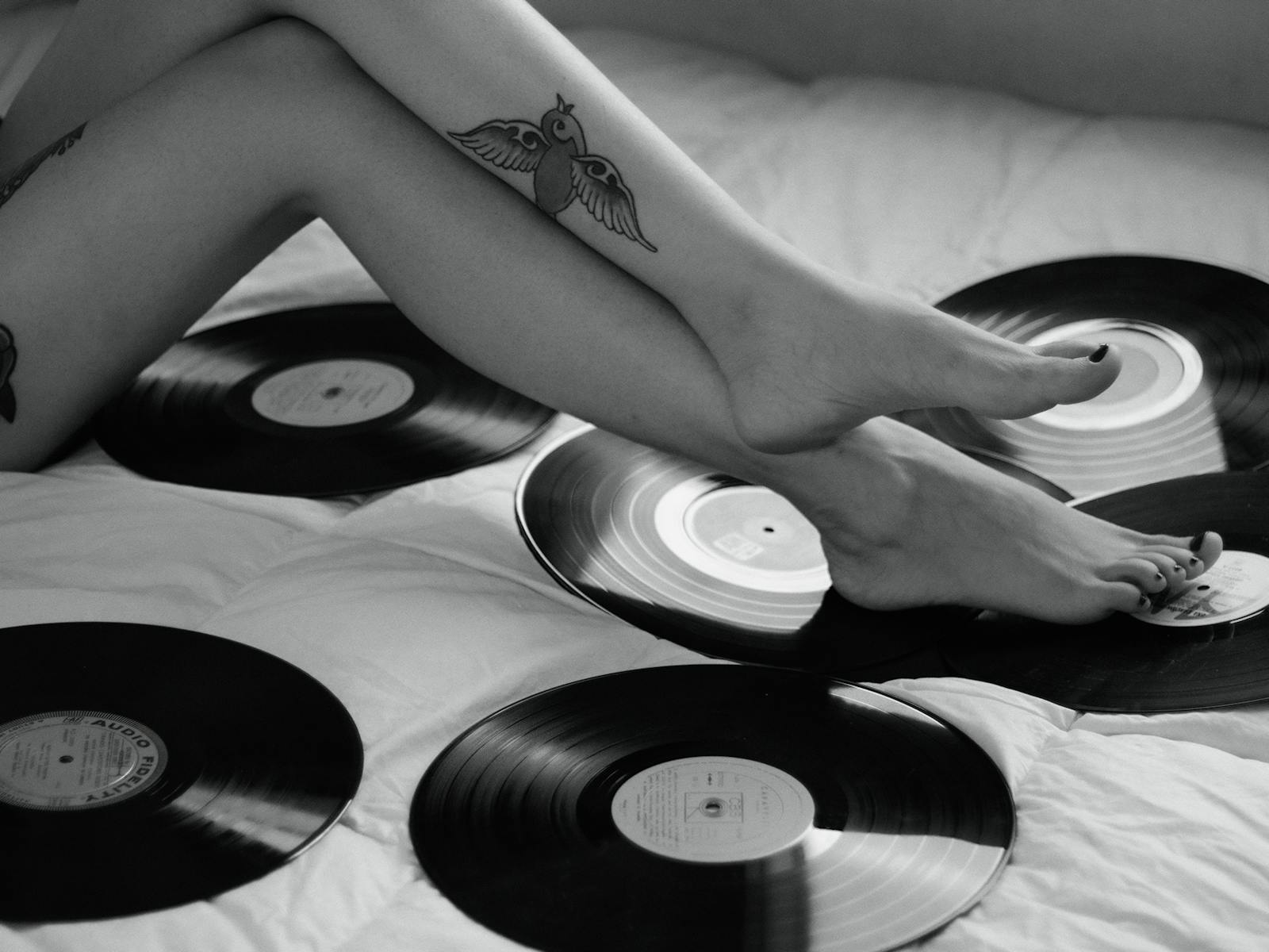 Black and white photo featuring tattooed legs surrounded by vinyl records on a bed.
