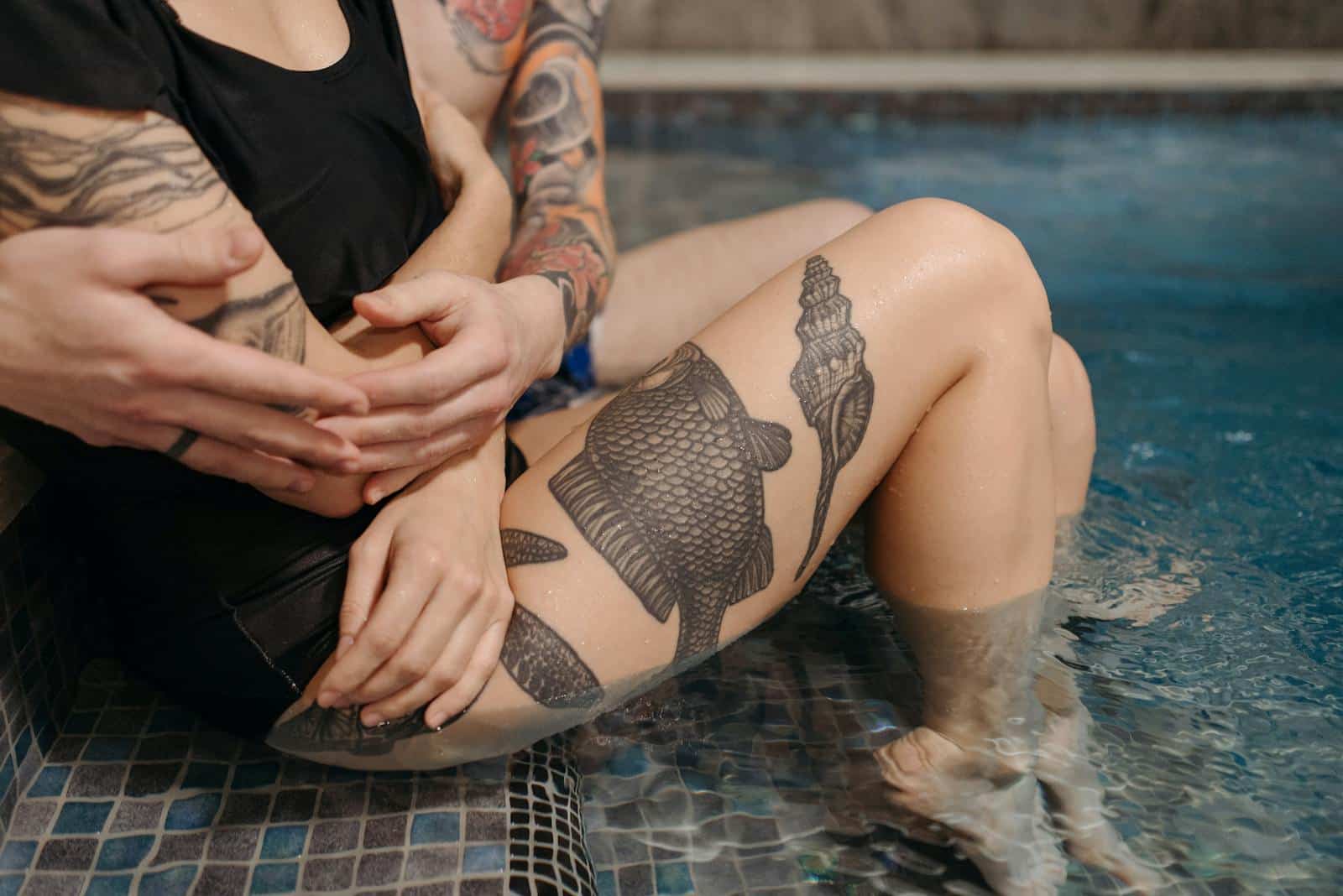A tattooed couple shares an intimate embrace while sitting by a swimming pool.