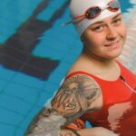Tattooed female swimmer in red suit posing in an indoor pool, exuding confidence and style.