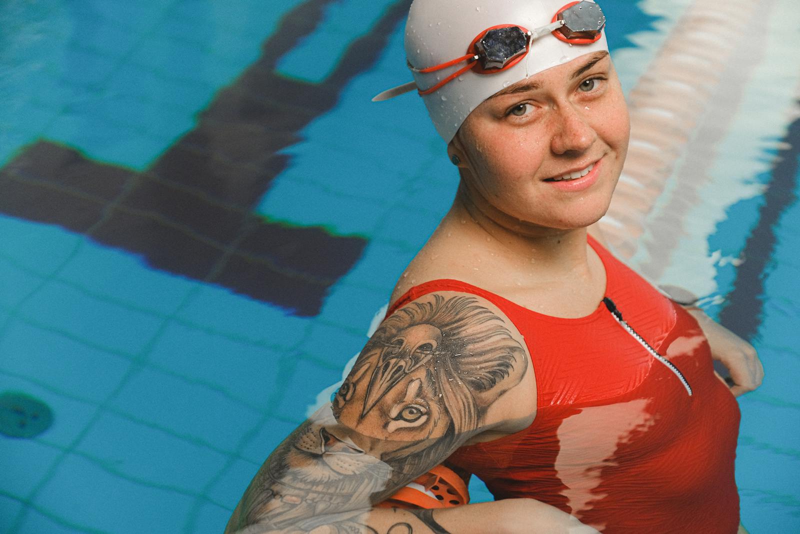Tattooed female swimmer in red suit posing in an indoor pool, exuding confidence and style.