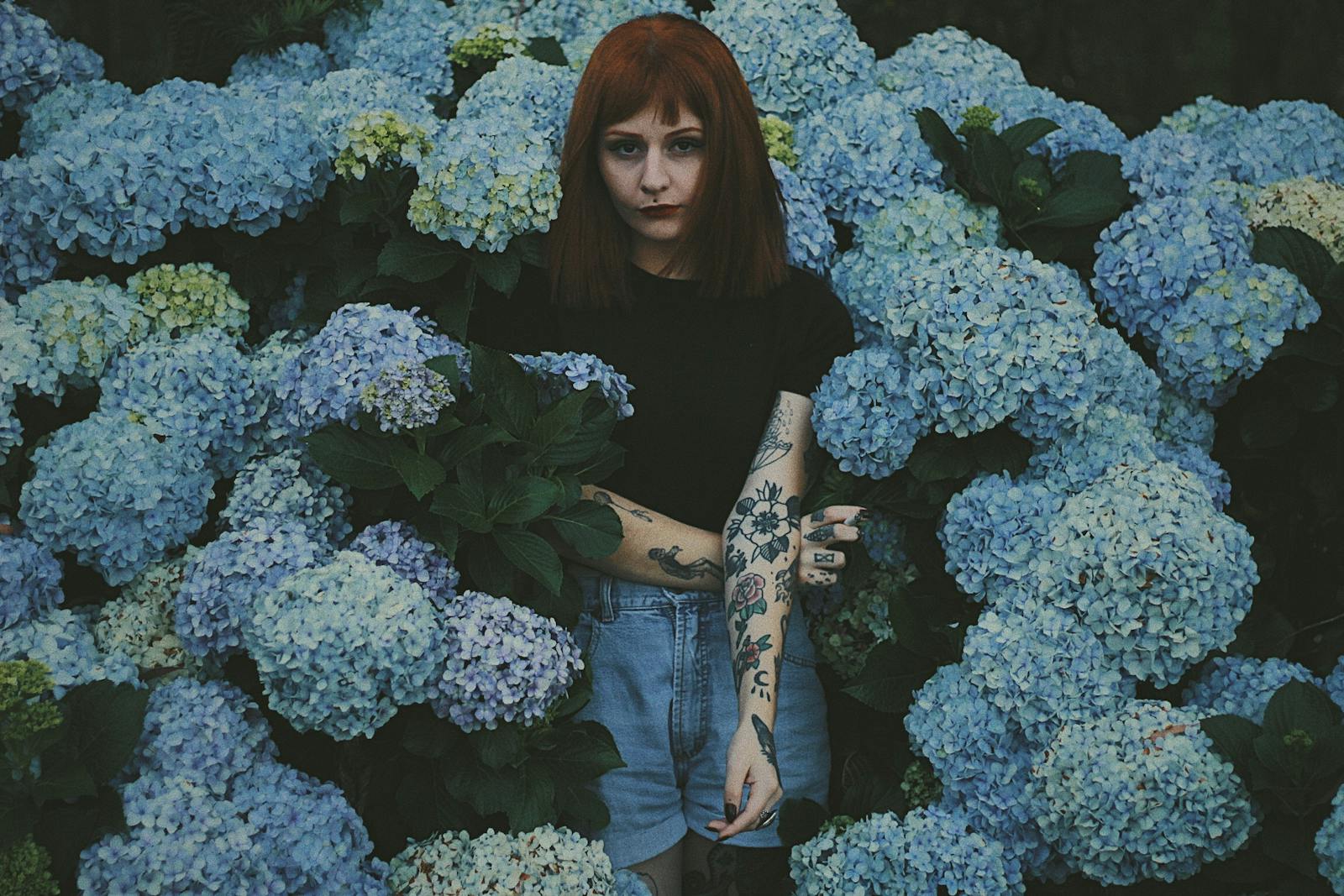 Artistic portrait of a woman standing amidst vibrant blue hydrangeas, conveying a moody and tattooed aesthetic.